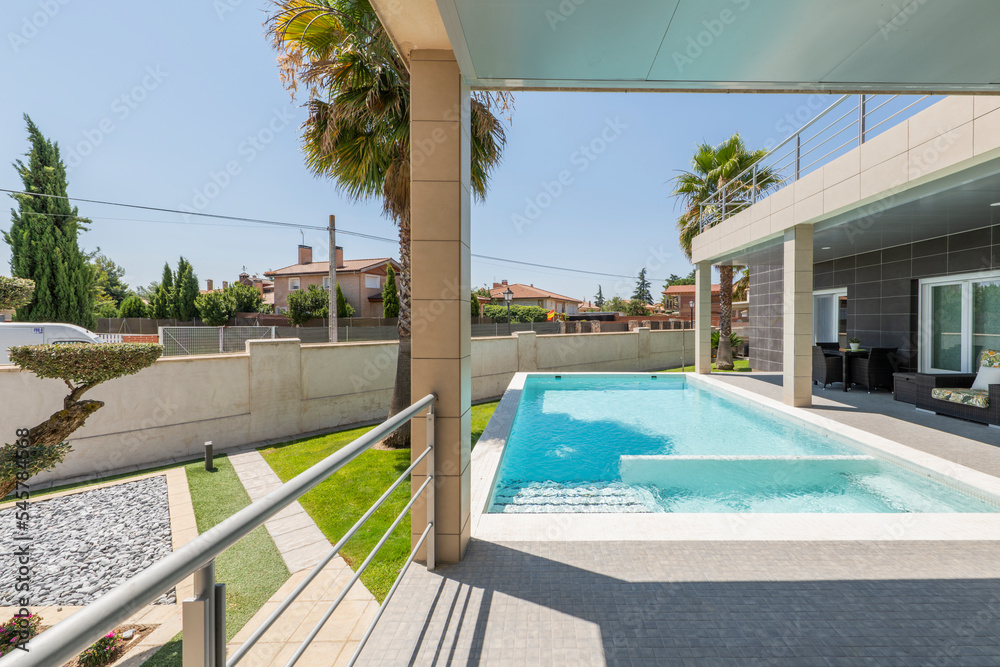 Porch of a single-family home with a swimming pool and palm trees, a metal railing overlooking the garden and shady wicker furniture