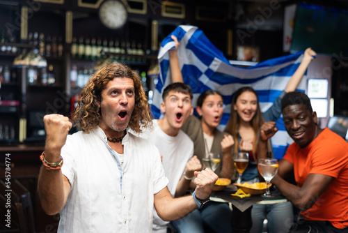Happy multiracial soccer fans with flag of Greece celebrating victory of team with pint of beer and chips in the pub