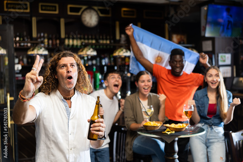 Excited adult man cheering for favorite team while watching match in sports bar on background of friends holding national flag of Argentina