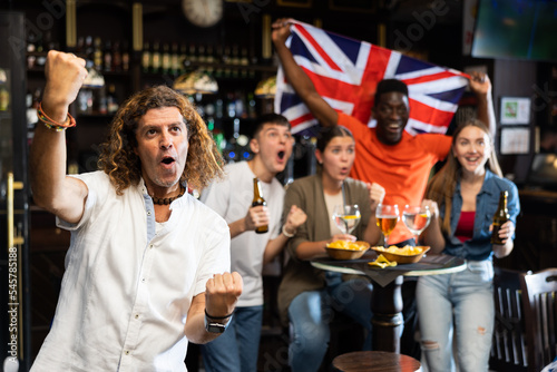 Excited diverse soccer supporters with flag of Great Britain celebrating victory with pint of beer and chips in the pub