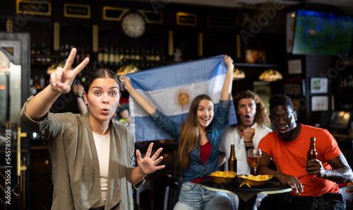 Excited young woman, Argentina football team fan, spending time in bar with friends, making victory sign with fingers. People with state flag in pub.