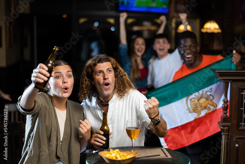 Man and woman, Iran football team fans, spending time in bar, drinking bear and screaming chants. People with state flag in pub.