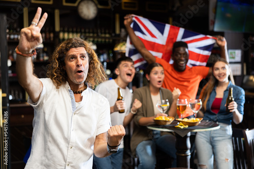 Male United Kingdom football team fan spending time in bar, making victory sign with fingers and screaming chants. People with state flag in pub.