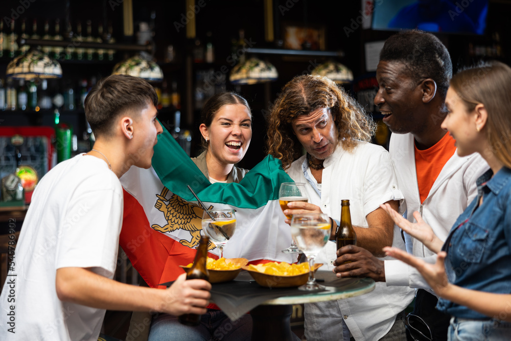 Cheerful multiracial soccer fans waving the flag of Iran while drinking beer and eating chips in sport bar