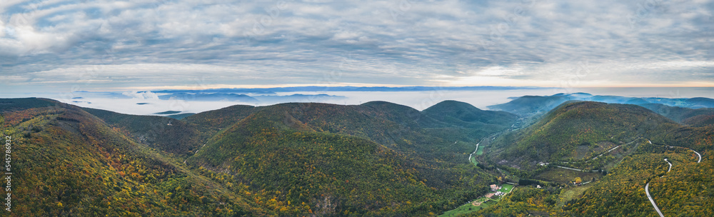 Panoramic view of the mountains covered with forests in the autumn aura. Autumn over the mountains with a thick morning fog in the background. A winding mountain road. In the vicinity of Mirmande