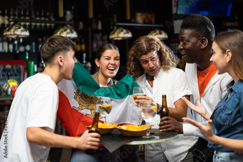 Cheerful multiracial soccer fans waving the flag of Iran while drinking beer and eating chips in sport bar