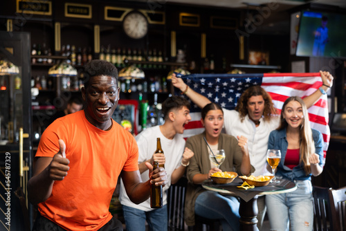 Happy excited african american man watching baseball in sports bar, giving thumbs up approving good game of favorite team against background of fans waving USA national flag..