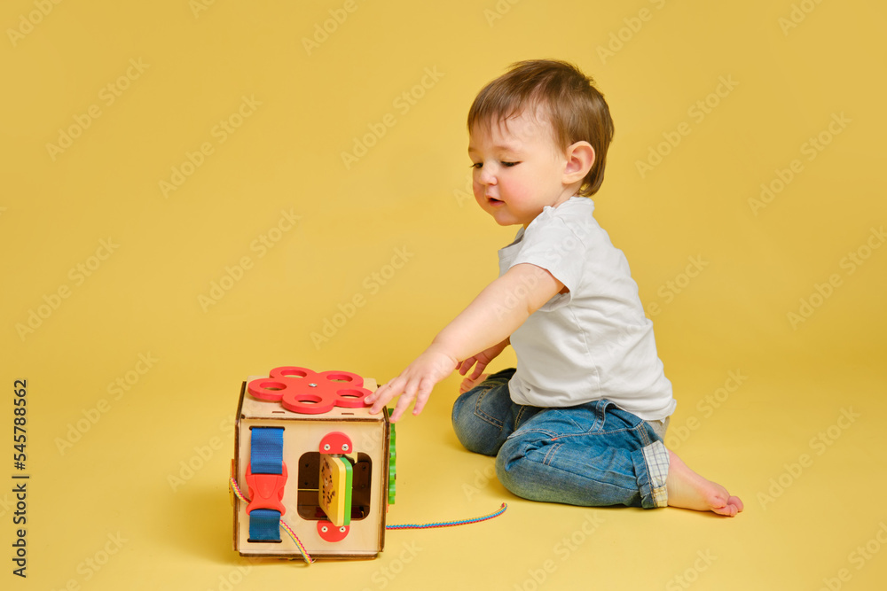 Toddler baby is playing logic educational games busyboard on a studio yellow background. Happy child play with educational toy, learning logic, copy space. Kid aged one year and four months Toddler baby is playing logic educational games busyboard on a studio yellow background. Happy child play with educational toy, learning logic, copy space. Kid aged one year and four months