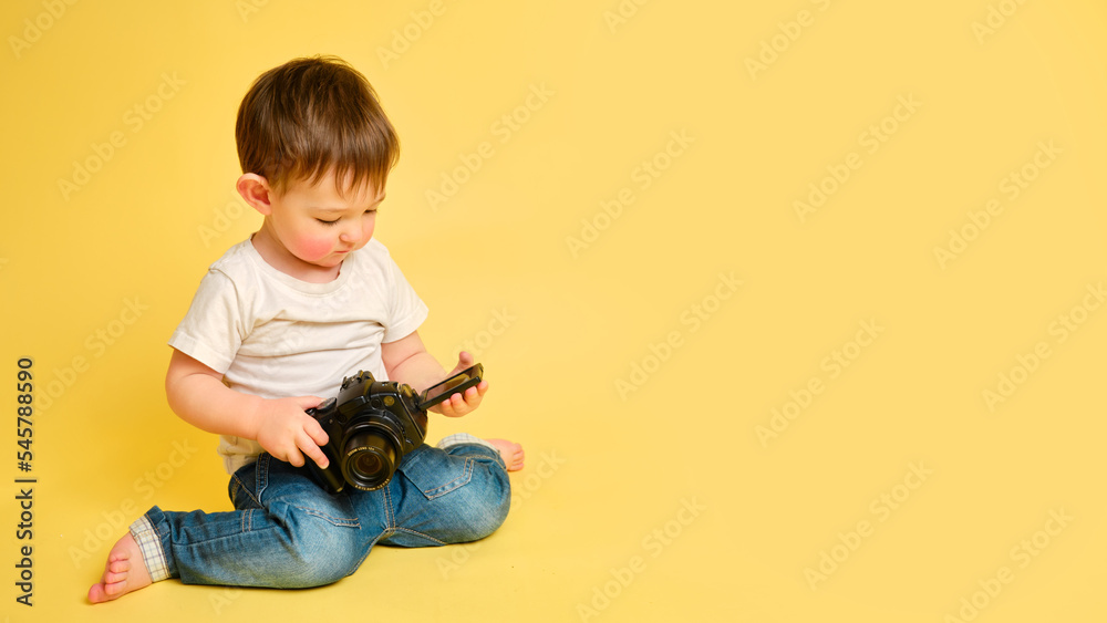 Toddler baby photographer with a camera on a studio yellow background. Happy child with photo equipment in a white t-shirt and blue jeans, copy space. Kid aged one year and four months Toddler baby photographer with a camera on a studio yellow background. Happy child with photo equipment in a white t-shirt and blue jeans, copy space. Kid aged one year and four months