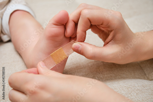 Mother woman sticks a medical band-aid on the toddler baby leg. Mom s hand with sticky wound protection tape and child s foot. Kid aged one year and three months