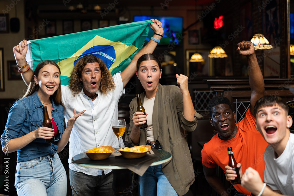 Fototapeta premium Emotional cheerful young adults friends, football fans cheering for favorite Brazil team together while watching match on tv in sports bar