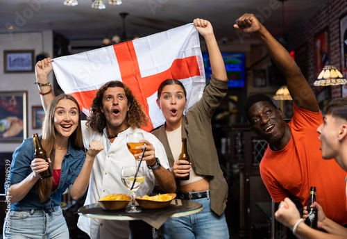 Excited young adult friends, sports fans celebrating victory of favorite football team with beer in bar, waving national flag of England