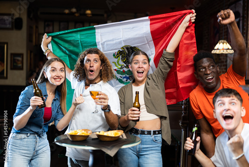 Multiracial Mexico sports fans, men and women, supporting their favourite team in bar, raising state flag and screaming chants together.
