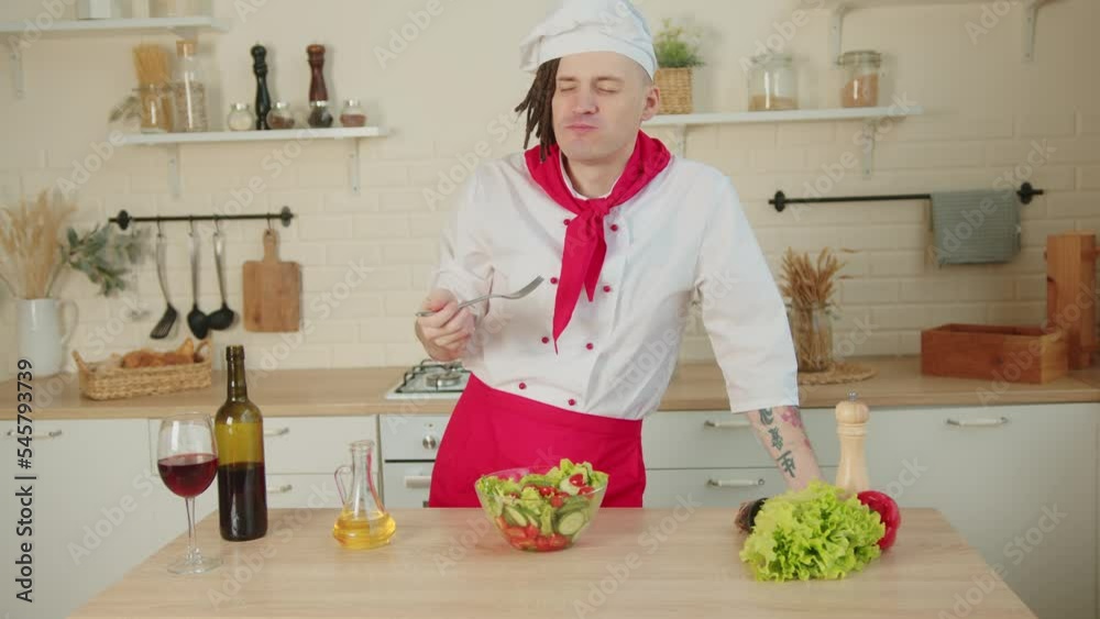 Young chef with dreadlocks eating vegetable salad in kitchen. Hungry man in uniform eating healthy vegetables. Young chef with dreadlocks eating vegetable salad in kitchen. Hungry man in uniform eating healthy vegetables.