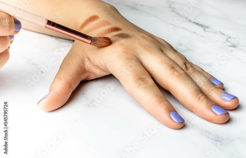 Hands of a Caucasian woman testing makeup foundation shades on her skin with a bristle brush. Darker earth tones to understand what works best in your daily skin care routine.