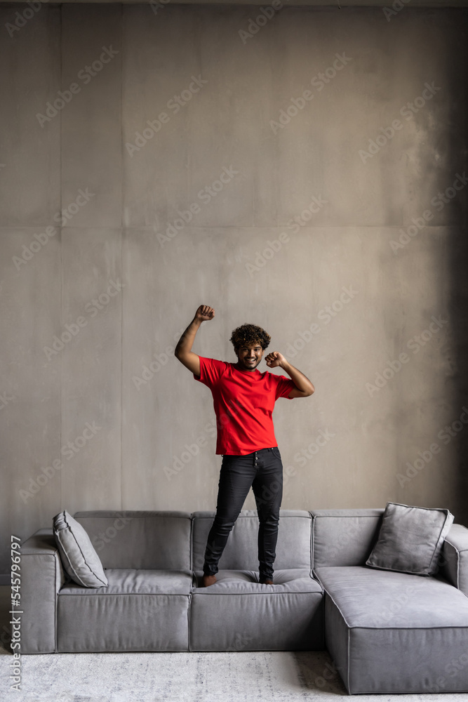 Young indian man having fun jumped on home couch listening to music on mobile phone with headphones dancing , singing and playing air guitar happy and crazy