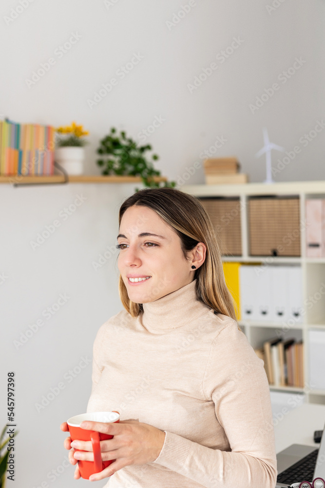 Vertical portrait of pensive young pretty woman holding a cup of coffee standing at home office looking away through the window Vertical portrait of pensive young pretty woman holding a cup of coffee standing at home office looking away through the window