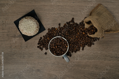 Top view of brown roasted coffee beans spilled on a table with a full coffee mug and biscuit