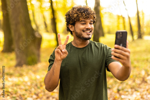 Young happy Indian man doing selfie pose on smart phone while standing in a park outdoor autumn, technology and communication