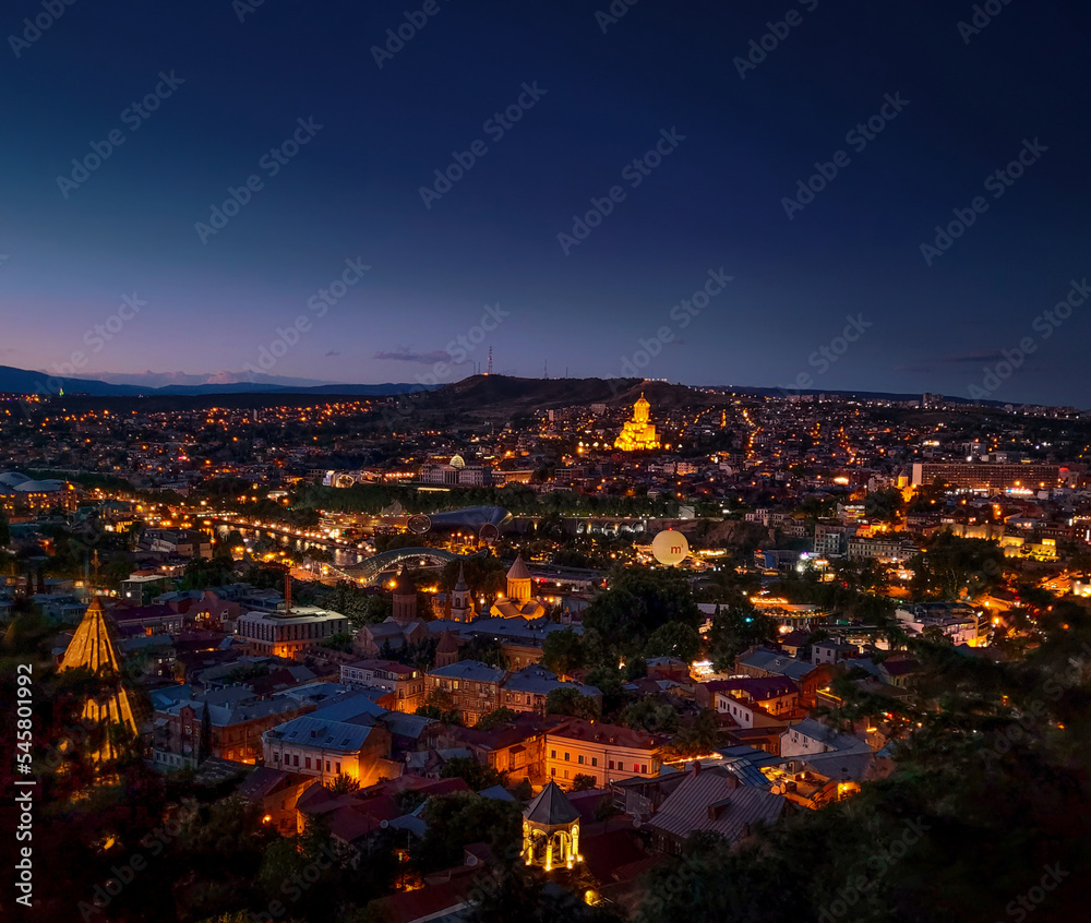 Summer night view on the city of Tbilisi Tiflis the capital of Georgia with streets of Old Tbilisi in foreground, Mtkvari Kura river, Holy Trinity Cathedral, Peace Bridge, State Palace of Ceremonies
