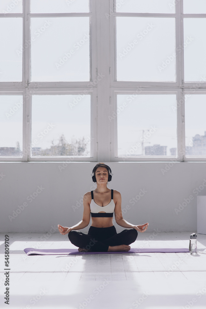 Confident young woman in headphones keeping eyes closed while meditating in front of the window