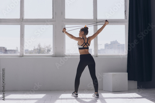 Rear view of fit young woman exercising with resistance band while standing in front of the window