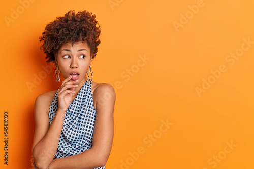 Studio shot of beautiful curly haired woman keeps mouth opened stares impressed at something awesome reacts to shocking news wears fashionable clothes isolated over orange background copy space