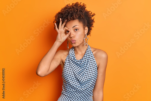 Pretty fashionable woman makes zero gesture over eye shows okay keeps lips folded being curious about something wears sleeveless polka dot blouse and earrings isolated over vivid orange wall