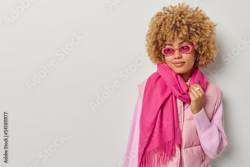 Thoughtful dreamy woman with curly hair looks pensively aside wears stylish sunglasses vest and scarf around neck concentrated aside isolated over grey background blank space for your promotion