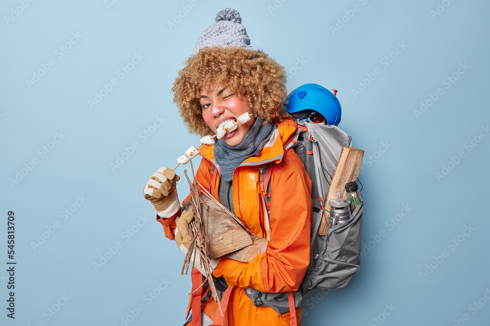 Horizontal shot of hungry female camper eats roasted marshmallow carries wood for making fire wears knitted hat and orange jacket poses with heavy rucksack on back isolated on blue background