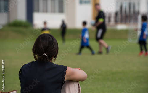 Mom sitting and cheering her son playing football in a school tournament on a sideline with a sunny day. Sport, outdoor active, lifestyle, happy family and soccer mom and soccer dad concept.