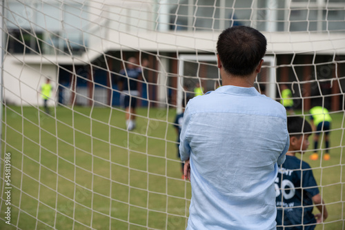 Dad standing and watching his son playing football in a school tournament on a sideline with a sunny day. Sport, outdoor active, lifestyle, happy family and soccer mom and soccer dad concept.