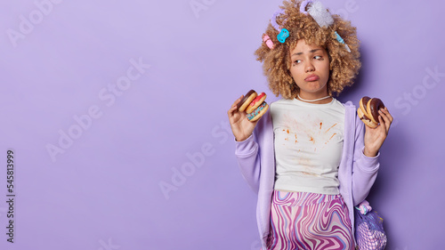 Sad expectant woman holds doughnuts eats sweet food has dirty clothes looks unhappily aside has bad mood isolated over purple background empty space for your promotional content. Expectations concept