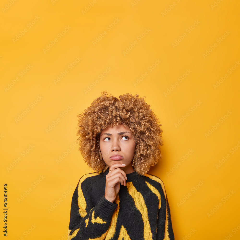 Thoughtful sad woman holds chin concentrated above with displeased expression feels frustrated wears casual jumper thinks about something unpleasant isolated over yellow background copy space Thoughtful sad woman holds chin concentrated above with displeased expression feels frustrated wears casual jumper thinks about something unpleasant isolated over yellow background copy space
