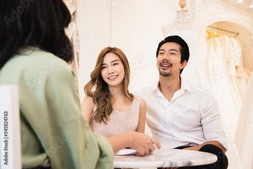 Smiling Asian woman wedding agent meeting with happy young husband and wife discussing contract, excited modern couple consider cooperation make deal with rental of dresses and weddings