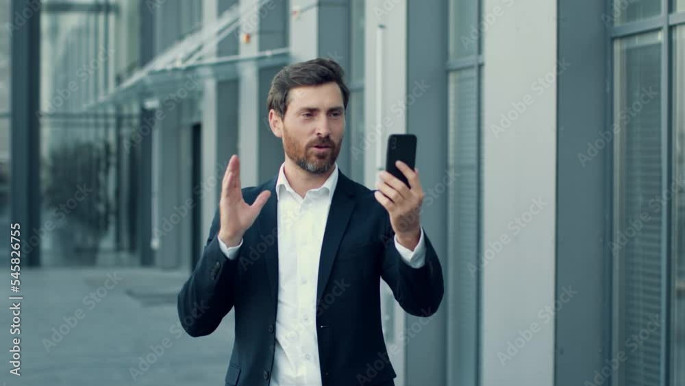 Follow shot of confident businessman video chatting with partners on go, walking near office building, free space Follow shot of confident businessman video chatting with partners on go, walking near office building, free space