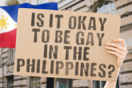 The question " Is it okay to be gay in the Philippines? " is on a banner in men's hands with blurred background. Friendly. Passionate. Contact. Date. Dating. Lover. Partner. Boyfriend. Pleasant. Appro