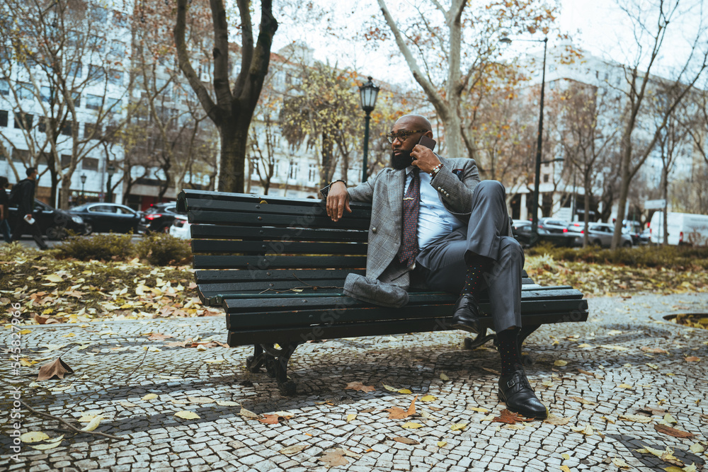 Fototapeta premium A wide-angle shot with a handsome mature bald African American man entrepreneur with a black beard, having a business conversation via his smartphone while sitting on a park bench on an autumn day