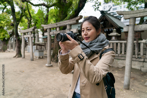 happy asian Chinese female photographer using digital camera to shoot pictures at Osaka Tenmangu Shrine in japan with several stone torii standing at background