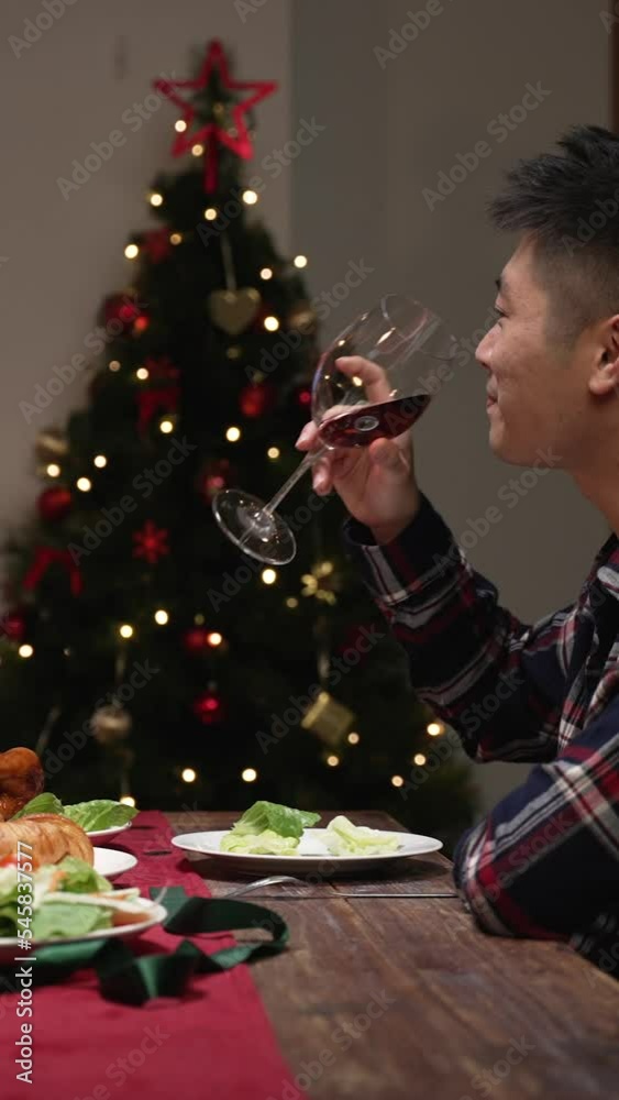 Vertical Screen: cheerful asian married couple chatting and drinking toast to celebrate new year while having cozy dinner date at home during with decorated Christmas tree Vertical Screen: cheerful asian married couple chatting and drinking toast to celebrate new year while having cozy dinner date at home during with decorated Christmas tree
