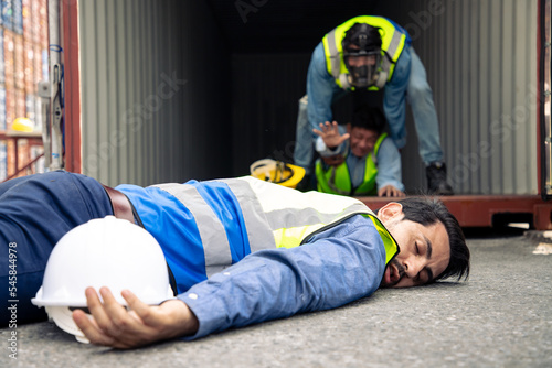 Handsome engineer lies unconscious on the ground after exposure to life-threatening chemicals. A container worker wearing a toxic mask is helping his friend.
