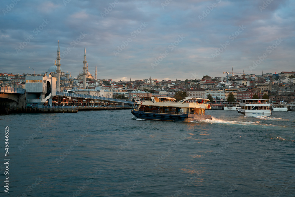 A large tourist boat floats on the river in the rays of the setting sun, against the backdrop of a Turkish city, with a beautiful mosque.
