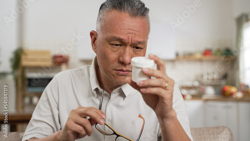 asian japanese old man with presbyopia holding and looking at a pill container with confusion. He squints his eyes trying to see the instruction on bottle clearly. eyes health issue problem