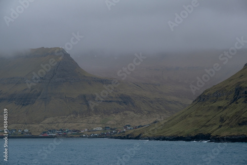 Impressive steep cliffs and rocky coastline silhouette coast of Faroe Islands in Atlantic Ocean on stormy grey day with clouds, lighthouse and hills seen from cruiseship cruise ship liner with spray