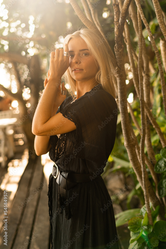 Fototapeta premium Close up portrait of pretty blond woman in boho stylish outfit posing outdoor in tropical garden. Stylish bohemian accessories.