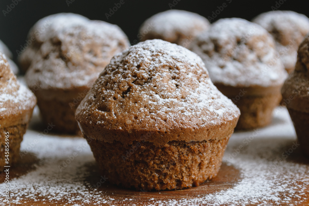 Close-up of a baked chocolate cupcake on a wooden board and sprinkled with white powdered sugar surrounded by other cupcakes