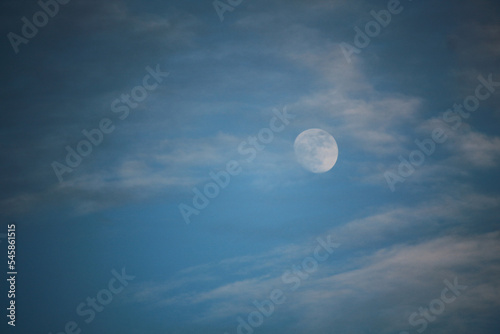 During the day, a beautiful moon rises in the blue sky in the middle of white clouds.