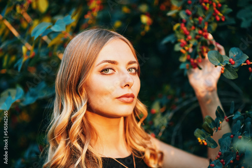 Young beautiful woman holds a branch with berries in her hand and looks at the camera. Outdoor portrait in the evening park