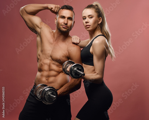 Fitness male and female models with equipment at pink background. Sporty couple with dumbbells in studio. Shirtless muscular man and slim girl with six pack abs posing with gym stuff