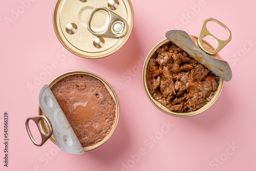 Wet cat food in open jars over pastel pink background. Wet pet feed in metal cans closeup. Canned meat pieces and soft pate for carnivore domestic animals. Сat food concept.